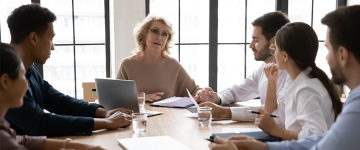 Six people sitting around a table having a business meeting looking at older woman talking.