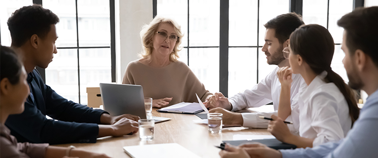 Six people sitting around a table having a business meeting looking at older woman talking.