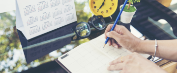 Person writing in planner with a blue pencil on a desk filled with a calendar, yellow clock, plant, and glasses.